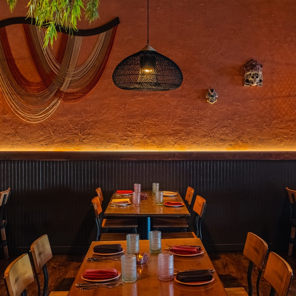 Restaurant interior with wooden tables set with plates and glasses, under a woven pendant light and textured orange wall.