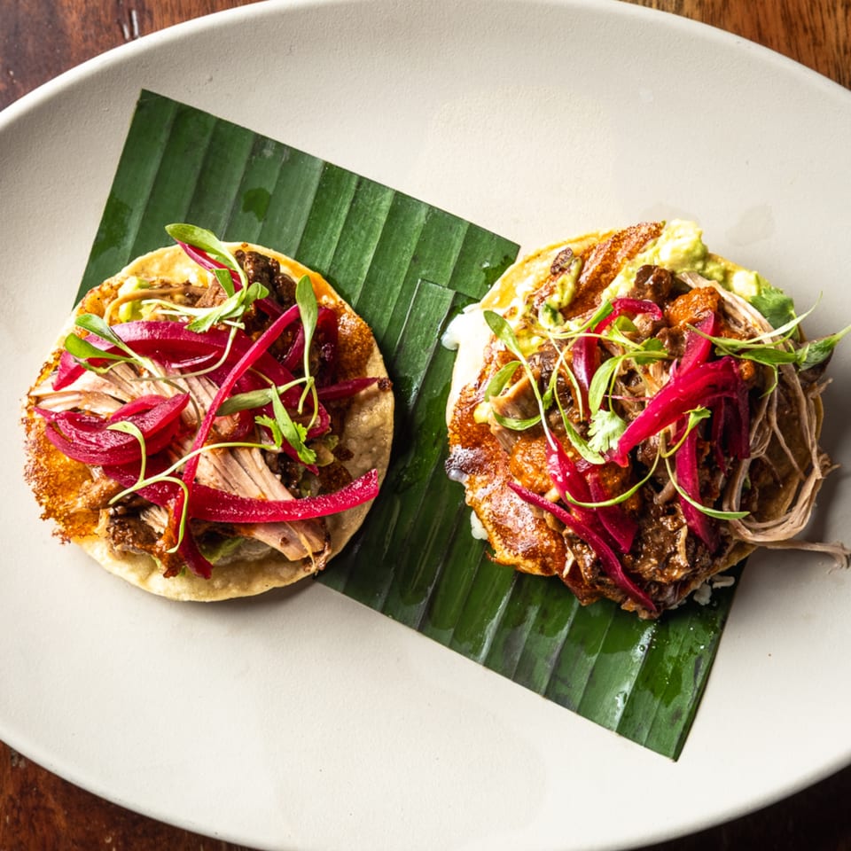Two La Casa De Juliette Mexican tacos with meat, red onions, and microgreens on a banana leaf.