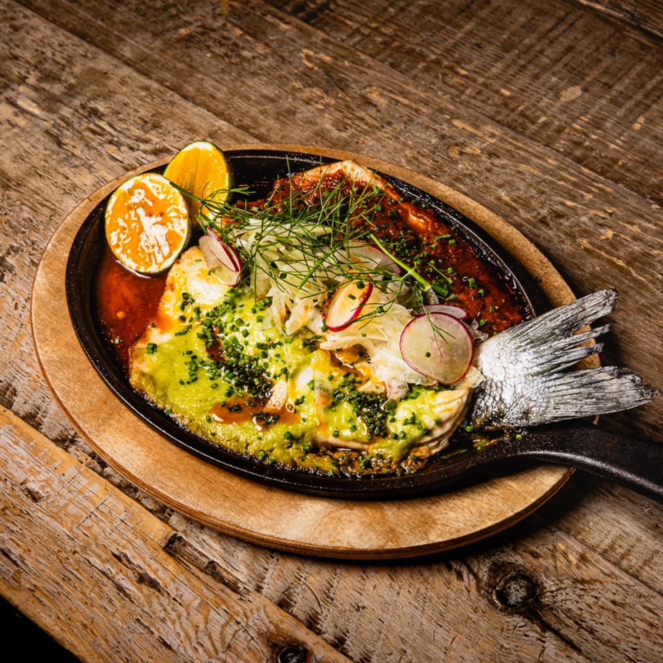 Whole grilled fish with green sauce, red sauce, herbs, radishes, and orange slices on a wooden table.
