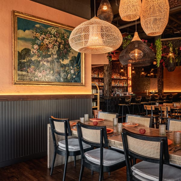 Restaurant dining room with a long wooden table, wicker chairs, and hanging woven light fixtures.