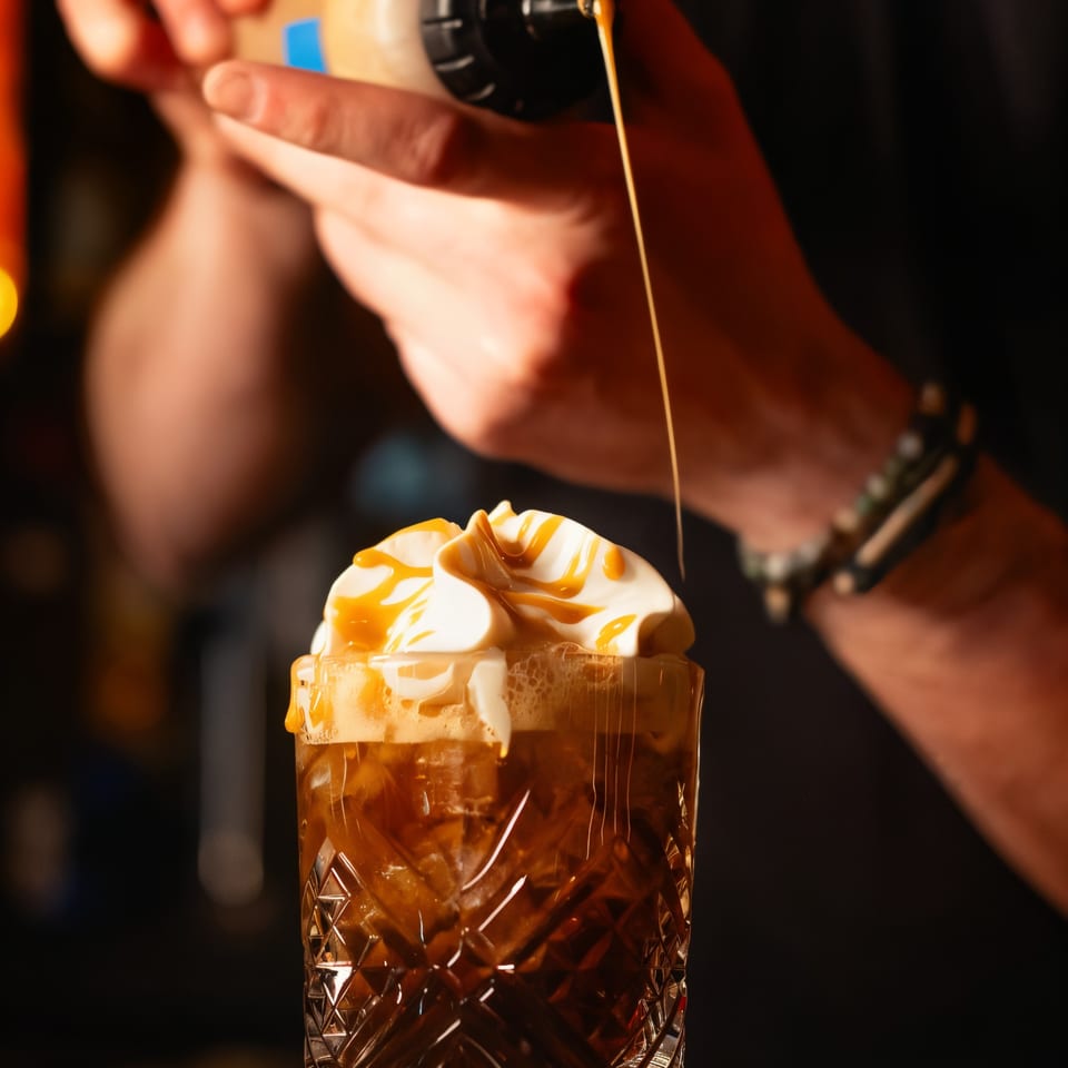 A bartender drizzles caramel sauce onto a drink topped with whipped cream in a textured glass.